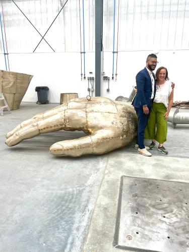 A man and a woman pose for a photo in front of a large golden sculpture of a hand.