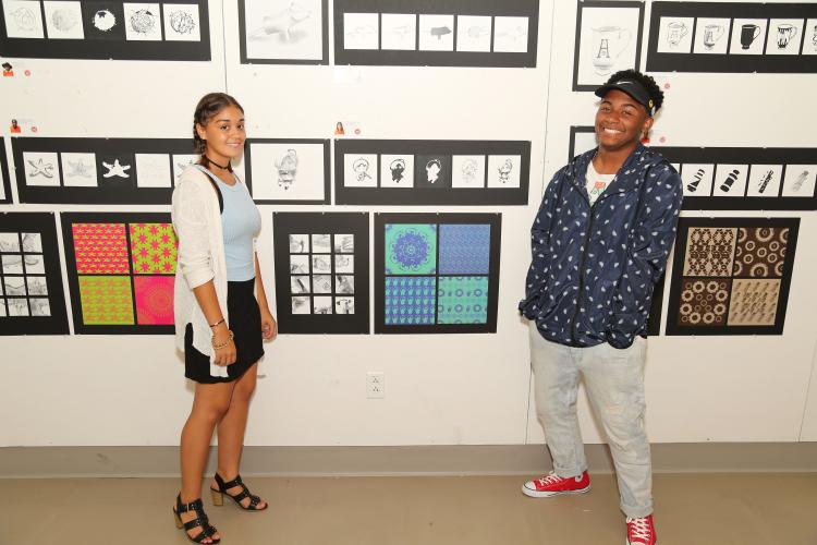 Two teenagers stand in front of a wall adorned with artwork