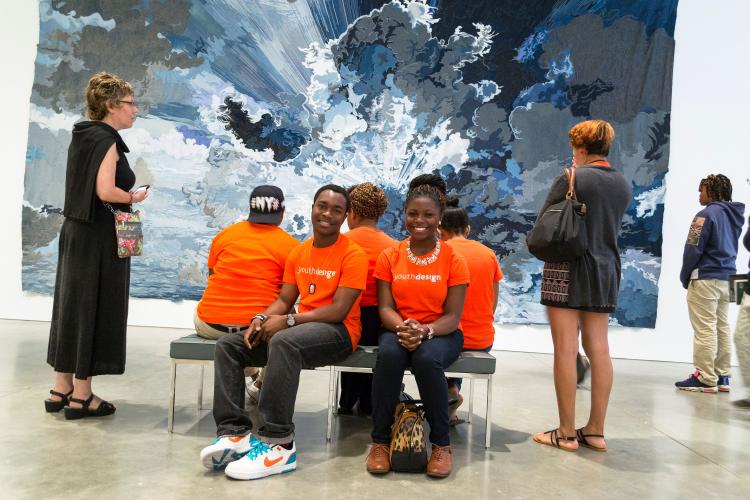 Five young adults dressed in bright orange T-shirts sit on benches in front of a large artwork hanging on a wall